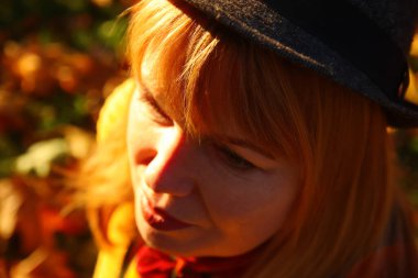 Defocus closeup portrait of candid young Caucasian blonde woman in colorful autumn park. Bright dark stylish woman in hat and shawl of neck on fall background. Happy dreaming concept. Out of focus.