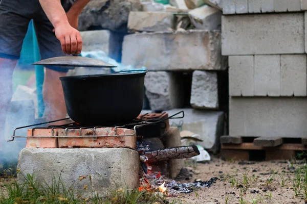 Defocus man cooking fish soup in the iron bowler over a campfire. Fish soup boils in cauldron. Soup in a pot in the fire. Camp at day. Nature green grass background. Out of focus