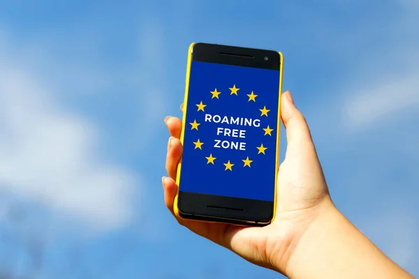 Roaming Free Euro Zone. Woman hand holding black smartphone in yellow plastic case on blue sky nature background, sunny day. Black screen. Female hand. Smart phone. European Union flag.