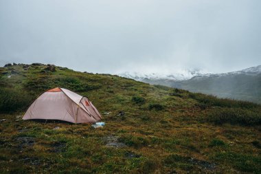 Yeşil tepelerde kayaların arasında çadırları olan manzaralı bir dağ manzarası. Alçak bulutlarda kar kaplı güzel dağlar. Dağlık bölgelerde, bulutlu havalarda çadırlı atmosferik manzara. Yüksek dağlar karla kaplı.
