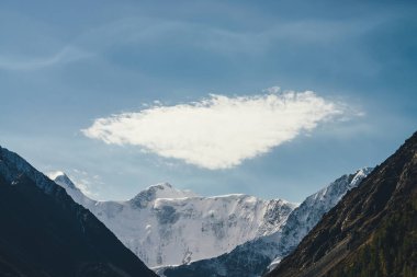 Karla kaplı yüksek dağlı, sirrus bulutlarının altında tepesi karla kaplı minimalist dağ manzarası. Bulutlu gökyüzünde kar beyazı, keskin zirvesi olan güzel bir dağ manzarası. Harika dağlar..