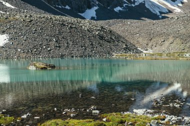 Turkuaz dağ gölü, şeffaf su ve taştan tabanlı güzel manzara manzarası. Güneş ışığında berrak su yüzeyi olan gök mavisi bir buzul gölü. Dağ gölü yakınlarında beyaz kar ve yeşil çimen.