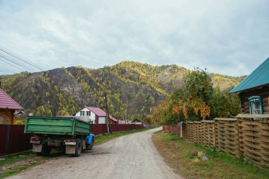 Sonbahar renklerinde canlı bir manzara. Sonbahar ormanı olan dağlara bakan kır evleri arasında köy çitlerinin kenarına park etmiş mavi yeşil bir Rus kamyonu var. Rusya, Altai Cumhuriyeti, 19 Eylül 2020.