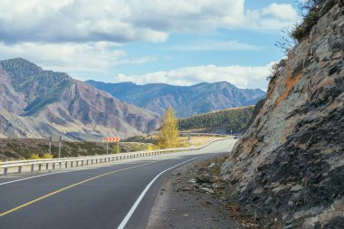 Renkli sonbahar manzarası, gün ışığında kayalar boyunca uzanan dağ yolu. Sonbahar renklerinde dağ yolu olan parlak alp manzarası. Sonbahar zamanı dağlarda otoyol. Yol boyunca güzel kayalık duvar boyunca.