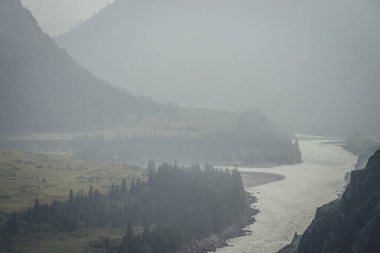 Geniş dağ nehri olan puslu dağ manzarası. Sis içindeki iki dağ nehrinin birleştiği koyu yeşil kasvetli manzara. Büyük nehirlerin yağmurlu havada birleşmesine karanlık atmosfer manzarası.