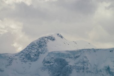Bulutlu gökyüzünün altında yüksek karlı dağ duvarı olan atmosferik dağ manzarası. Karla kaplı, bulutlu bir havada, dağların tepesinde dramatik bir manzara. Kar tepesinde bulutların arasından gün ışığı.