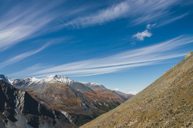 Sonbahar renklerinde büyük dağları olan renkli alp manzarası. Mavi gökyüzünde, sirrus bulutlarının altında, güneş tepede karla kaplı. Güneşli kayalar ve karla kaplı dağ tepeleri ile resimli sonbahar manzarası