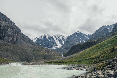 Bulutlu gökyüzünün altındaki karlı dağların arka planında dağ gölü olan güzel bir manzara. Buzul gölü ve yüksek dağlı buzullu atmosferik yeşil manzara. Müthiş Highland Manzarası.