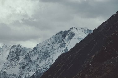 Bulutlarda karlı dağ duvarları olan atmosferik bir manzara. Yüksek karla kaplı, bulutlu gökyüzünün altında soluk tonlarda, kasvetli dağ manzarası. Karlı sivri tepeler ve koyu renkli kayalar..