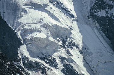 Beyaz buzullu ve kar tanecikli büyük kayalık dağ duvarının doğal arka planı. Güneşli karlı dağ yamacının güzel doğal dokusu. Siyah beyaz yüksek dağlardan oluşan bir çerçeve.