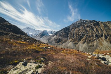 Sonbahar güneşli manzarası Highland Valley ve karla kaplı yüksek dağlar sirrus bulutlu mavi bulutlu gökyüzü altında. Sonbaharda kayalık dağların arasında güneşli vadiyle atmosferik dağ manzarası.