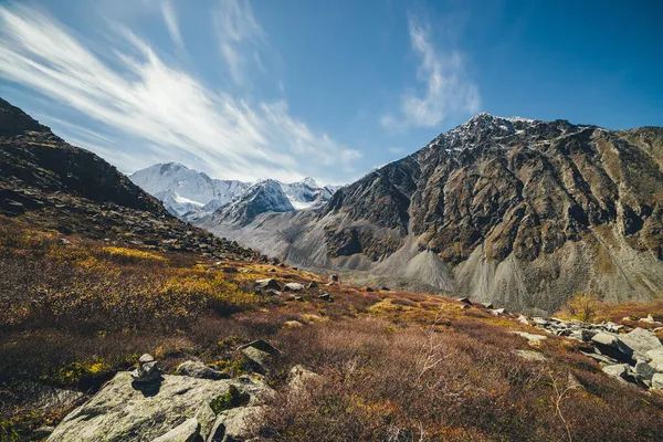 Autumn sunny landscape with highland valley and snow-covered high mountains under blue cloudy sky with cirrus clouds. Atmospheric mountain scenery with sunlit valley among rocky mountains in autumn.