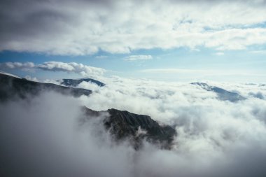 Yoğun alçak bulutlarda büyük kayalar ve dağlar olan harika bir dağ manzarası. Atmosferik dağlık arazide bulutların tepeleri var. Yoğun bulutların üzerinde kar dağlarının güzel manzarası.
