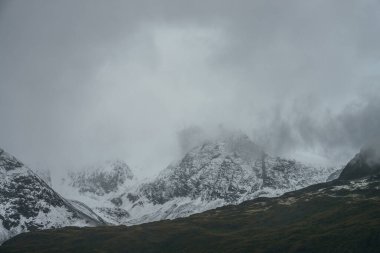 Minimalist kasvetli dağlık arazi karla kaplı yüksek dağlar ve bulutlu hava. Alçak bulutlarda karlı dağ sıraları. Atmosferik minimalizm. Yeşil dağlar, beyaz kar ve gri gökyüzü..