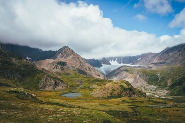 Güneşli yeşil vadide dağ gölü ve bulutlu gökyüzünün altında buzul manzarası. Muhteşem güneşli manzara, güzel buzul gölü, güneş ışığı altında alçak bulutlarda büyük dağ sıralarına karşı.