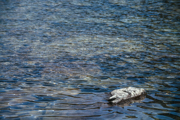 Природный фон с камнем в голубой чистой воде горного озера. Живописный природный фон рябь на голубой водной поверхности ледникового озера. Минимальный фрагмент ледникового озера с камнем.