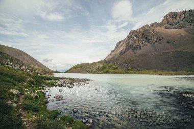 Bulutlu gökyüzünün altında, ufuktaki dağların arkasındaki dağ gölüne güzel bir manzara. Yüksek dağ vadisinde gölü olan atmosferik yeşil bir manzara. Dağlık vadide dağ gölü manzarası