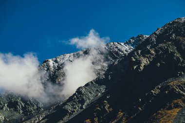 Atmosferik dağ manzarası, yüksek siyah kayalık dağ karlı ve alçak bulutlu dağ. Alp manzaralı, alçak sirrus bulutlu, büyük, karanlık bir dağ manzarası. Kayalıklarda kar ve bulutlar.