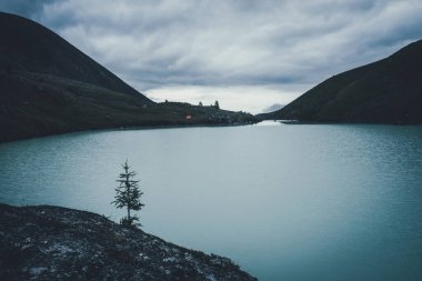 Alacakaranlıkta dağ gölü suyuna karşı köknar. Karanlık zamanlarda bulutlu gökyüzünün altındaki dağlık vadide turuncu çadırı olan karanlık bir yer. Dağlık göle karşı tek ağaçlı karanlık bir manzara..