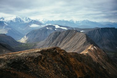 Dağ vadisinin arka planında keskin kayalık tepeleri ve bulutlu gökyüzünün altında buzulları olan karlı dağ sıraları olan muhteşem dağlık arazi. Keskin kayalara ve büyük karlı dağlara manzaralı hava manzarası.
