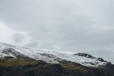 Bulutlu gökyüzünün altında kar kaplı dağlarla minimalist dağlık arazi. Bulutlu havada karlı yüksek dağ sıraları. Atmosferik minimalizm. Yeşil dağlar, beyaz kar ve gri gökyüzü..
