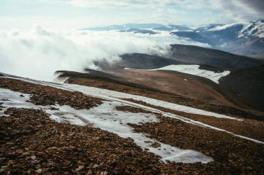 Yoğun alçak bulutlar altında ufukta dağlardan oluşan manzaralı dağlık bir arazi. Dağlık bölgelerdeki bulutlarda harika katmanlı dağ rahatlaması. Karlı dağlar ve yüksek irtifada kalın bulutlarda buzullar.