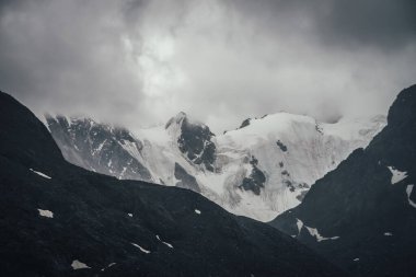 Karanlık atmosferik dağ manzarası kurşunlu gri bulutlu gökyüzünde kara kayalar üzerindeki buzullar. Yağmurlu havada, alçak bulutlarda, karlı dağlar. Kara kayalık dağların sisli olduğu kasvetli bir manzara..