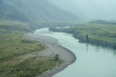 Geniş dağ nehri olan güzel sisli dağ manzarası. Koyu yeşil kasvetli manzara ve sisli dağ nehri. Yağmurlu havada, büyük dağların arasındaki büyük nehrin karanlık atmosferik görüntüsü.