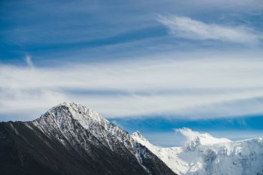 Atmosferik alp manzarası. Mavi gökyüzünde, sirrus bulutlarının altında karla kaplı dağ tepesi. Karla kaplı sivri uçlu güzel dağ manzarası ve alçak bulutlu yüksek karlı dağ duvarı..