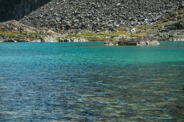 Turkuaz dağ gölü olan güzel bir manzara. Güneş ışığında gök mavisi bir buzul gölü. Dağ gölünün berrak su yüzeyine sahip renkli güneşli bir manzara. Şeffaf su ve taştan tabanlı buzul gölü.