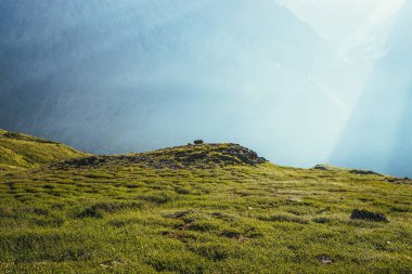 Arkasında kayalar ve tepeler olan renkli yeşil bir manzara. Güneş ışığıyla kaplı dev bir dağ duvarı. Güneş ışınları ve güneş patlamalarıyla minimalist, canlı, güneşli manzara. Minimum alp manzaralı. Sahne minimalizmi.