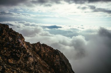 Bulutlu gökyüzünün üstündeki dağın arka planında keskin kayalar. Dağ tepesi kalın bulutlarda yüzer. Yoğun alçak bulutların üstünde dağ tepesi olan minimalist bir manzara. Yoğun bulutlar üzerinde sahne minimalizmi.