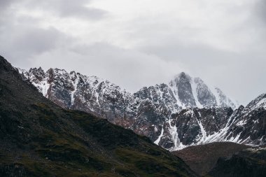 Atmosferik dağ manzarası alçak bulutlarda karla kaplı. Bulutlu havada keskin zirvesi olan dramatik dağ manzarası. Bulutlu gökyüzünde karlı, sivri tepeye müthiş bir manzara.