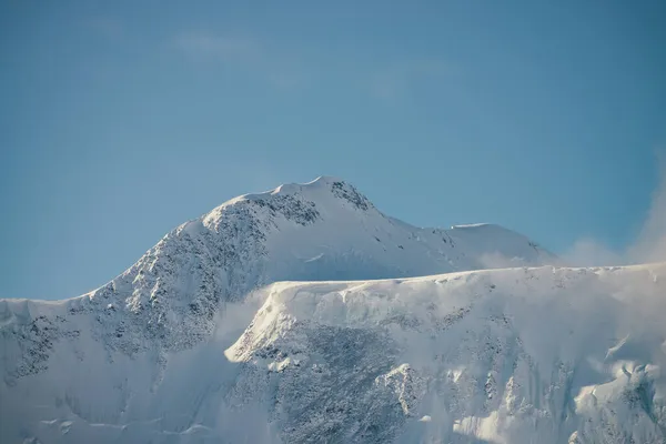雪のミニマリストビュー帽をかぶった太陽の下で厚い低い雲の中の山の壁 青空に濃い雲の間に白い雪のピークと風景明るい山の風景 雪の上で素晴らしい景色 — ストック写真