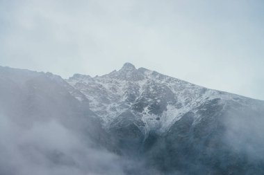 Siste sivri uçlu dağ siluetine harika bir manzara. Atmosferik sisli dağ manzarası, karla kaplı ve alçak bulutlarda zirveye ulaşmış. Sisli kayalık tepeli minimalist alp manzarası