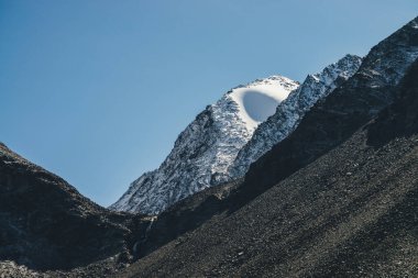 Mavi gökyüzünde siyah kayaların üzerinde beyaz karla muhteşem bir dağ manzarası. Atmosferik alp manzarası, kar küreleri dağın tepesinde güneş ışığı altında. Güneş ışığının zirvesinde kar şeklindeki güzel sıradışı küre.