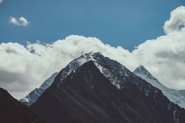 Atmosferik dağ manzarası. Karla kaplı büyük bir tepe noktası ve alçak bulutlarda koyu renkli karlı sivri bir tepe. Yüksek karlı, kasvetli dağ manzarası bulutlu gökyüzünün altında solgun tonlarda..