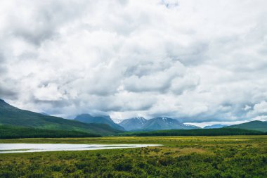 Dağ gölü ve ormanın olduğu dramatik bir manzara güneş ışığında tepeler ve değişken havada alçak bulutlarda karlı dağlar. Bulutlu gökyüzünde yüksek dağlar ve yeşil otların ve orman tepelerinin yakınında göl.