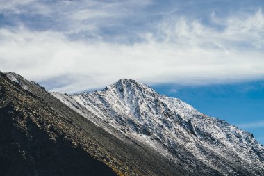 Mavi gökyüzünde, sirrus bulutlarının altında yüksek karlı dağ tepesi olan güneşli manzara. Güzel dağ manzaralı, harika, keskin tepeli, güneşli karlı. Atmosferik kayalık dağ tepesi güneş ışığında.