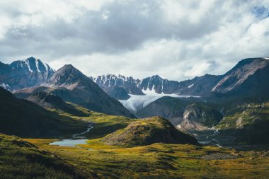 Atmosferik alp manzarası, yeşil vadide dağ gölü ve bulutlu gökyüzünün altında buzul. Güneşli tepeler ve dağ sırasına karşı kayalar arasında buzul gölü olan muhteşem dağlık arazi manzarası..