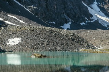 Turkuaz dağ gölünde yosunlu kayayla şeffaf su ve taşlı dipli güzel bir manzara. Güneş ışığında gök mavisi bir buzul gölü. Berrak su yüzeyinin üstünde yosunlu taşlar olan renkli bir manzara..