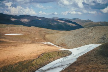 Güneşli dağ çöl ferahlığı, bulutlu gökyüzünün altında, yamaçta uzun bir buzul var. Güneşli manzaralı dağlık bölgede uzun küçük buzullar ve dağlarda eriyen su akıntıları..