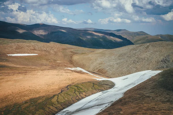 Sunny mountain desert relief with long glacier on slope in sunlight under cloudy sky. Sunny scenic highlands landscape with long small glacier on mountainside and melt water streams high in mountains.