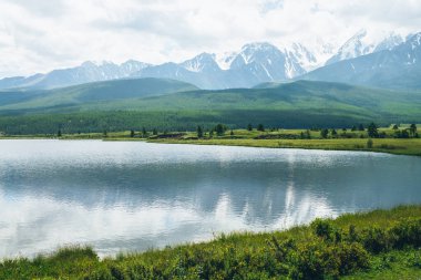 Dağ gölü ve ormanın olduğu dramatik bir manzara güneş ışığında tepeler ve değişken havada alçak bulutlarda karlı dağlar. Bulutlu gökyüzünde yüksek dağlar ve yeşil otların ve orman tepelerinin yakınında göl.