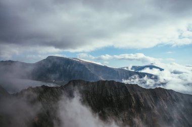 Büyük kayalar ve alçak bulutlarda dağlar olan harika sisli bir manzara. Atmosferik sisli manzara, dağ duvarı ve bulutlarda buzullar. Kayalık duvara ve bulutlardaki kar dağlarına güzel bir manzara.