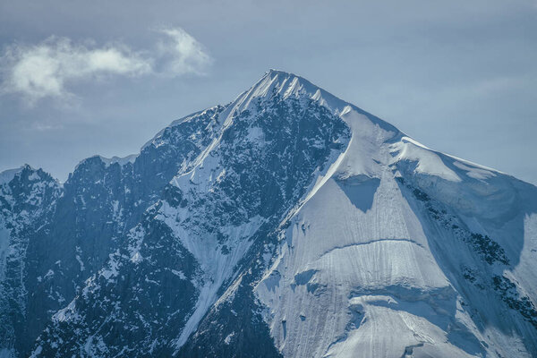 Awesome mountains landscape with big snowy mountain pinnacle in blue white colors and white cirrus clouds in blue sky. Atmospheric highland scenery with high mountain wall with pointed top with snow.