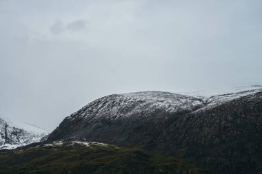 Bulutlu gökyüzünün altında kar kaplı dağlarla minimalist dağlık arazi. Bulutlu havada karlı yüksek dağ sıraları. Atmosferik minimalizm. Yeşil dağlar, beyaz kar ve gri gökyüzü..