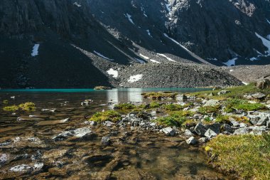 Turkuaz dağ gölü, şeffaf su ve taştan tabanlı güzel manzara manzarası. Güneş ışığında berrak su yüzeyi olan gök mavisi bir buzul gölü. Dağ gölü yakınlarında beyaz kar ve yeşil çimen.