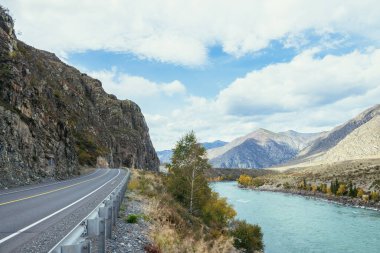 Parlak sonbahar manzarası, dağ yolu boyunca büyük dağ nehri boyunca güneş ışığı. Sonbahar renklerinde geniş turkuaz nehir ve dağ yolu olan parlak alp manzarası. Sonbahar zamanında dağlarda otoyol
