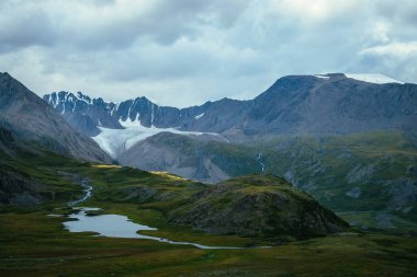 Atmosferik alp manzarası, yeşil vadide dağ gölü ve bulutlu gökyüzünün altında buzul. Güneşli tepeler ve dağ sırasına karşı kayalar arasında buzul gölü olan muhteşem dağlık arazi manzarası..